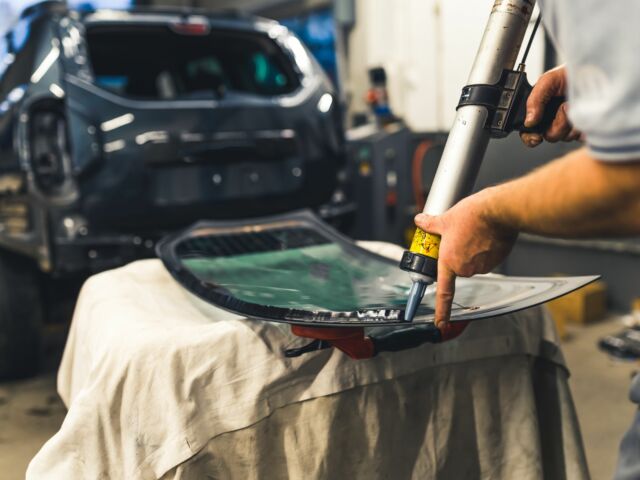 Installing process of car window. Indoor shot of professional unrecognizable mechanic using polyurethane glue gun. Black blurred car in the background.